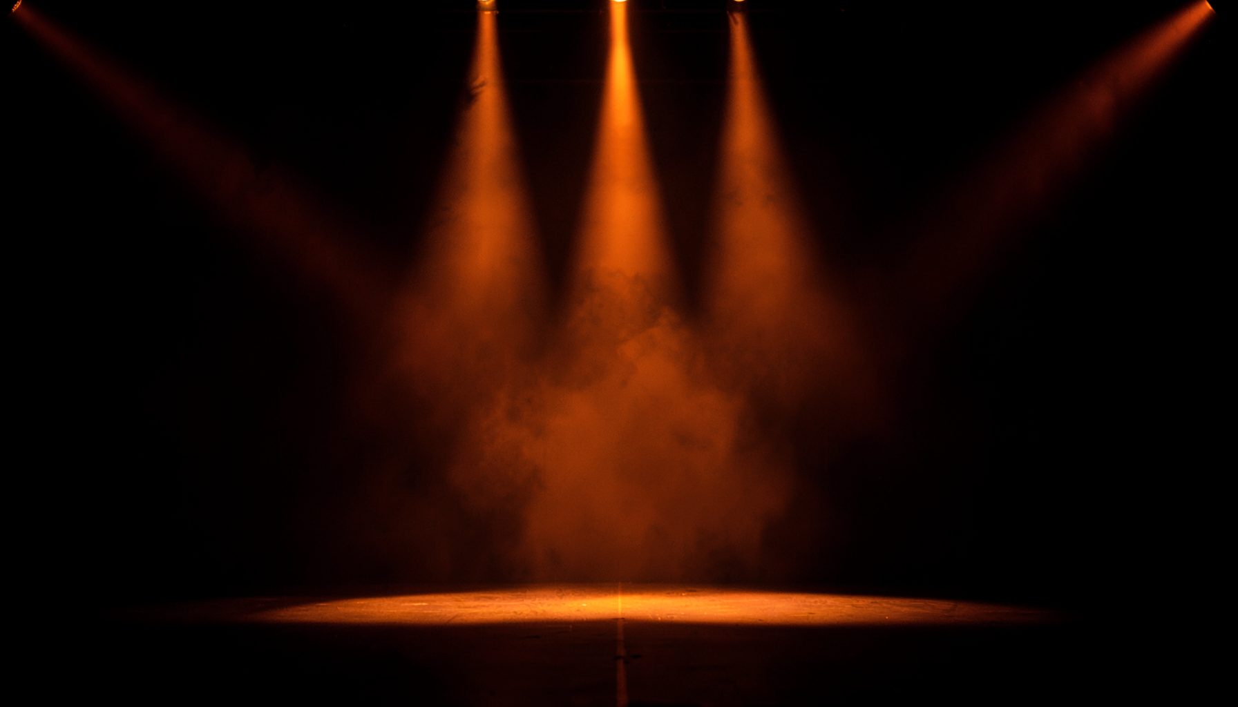 A symmetrical wide photograph of a stage with warm orange spotlight beams cutting through dark haze, deep black background