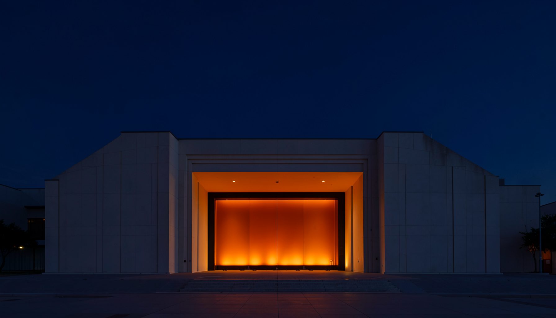 A wide symmetrical architectural photograph of a concert hall entrance at dusk, deep navy sky, warm orange stage lights